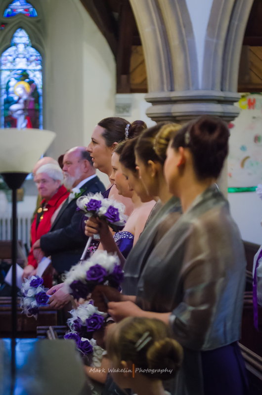 Bridesmaids and guests seated in a church during a wedding ceremony, holding purple and white flower bouquets.