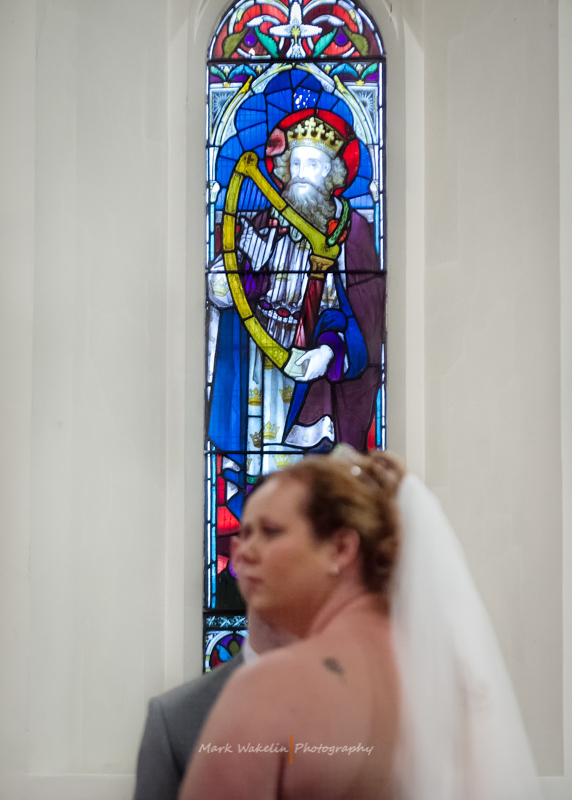 Stained glass window depicting a crowned king holding a harp behind a bride and groom in a church.