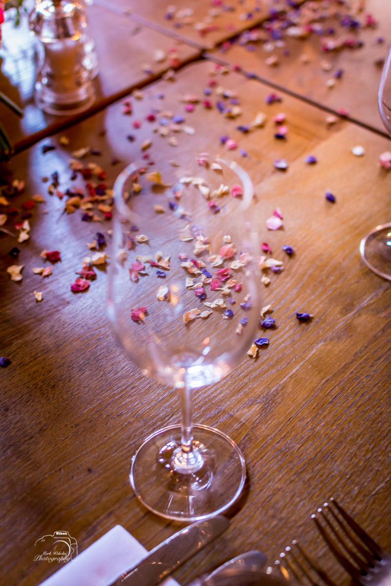 Empty wine glass on a wooden table scattered with colourful dried flower petals and set with cutlery.