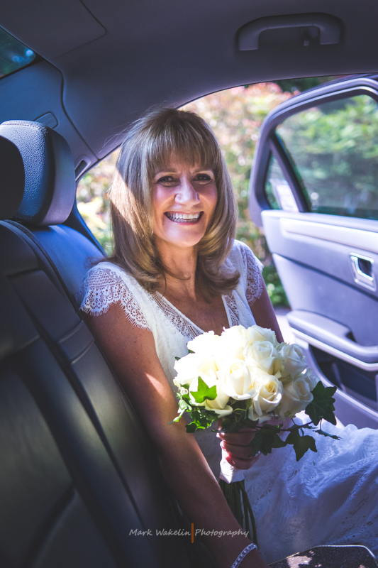 Smiling bride in a white lace dress holding a bouquet of white roses sitting in the backseat of a car with the door open.