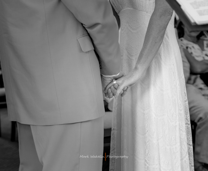 Black and white photo of a couple holding hands during a wedding ceremony, showing the bride's lace dress and the groom's suit.