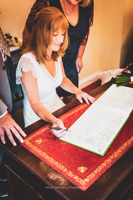 Bride in white dress signing a large book on a wooden table with a red cloth, with two people standing nearby.