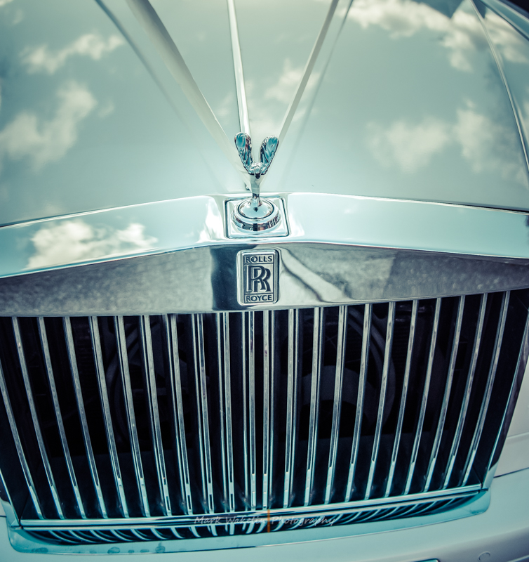 Close-up of a Rolls-Royce car grille and Spirit of Ecstasy hood ornament with sky reflections.