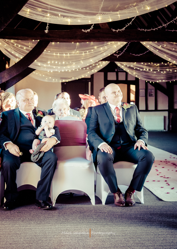Two men in suits seated in a decorated venue with one holding a baby, illuminated by draped fairy lights.