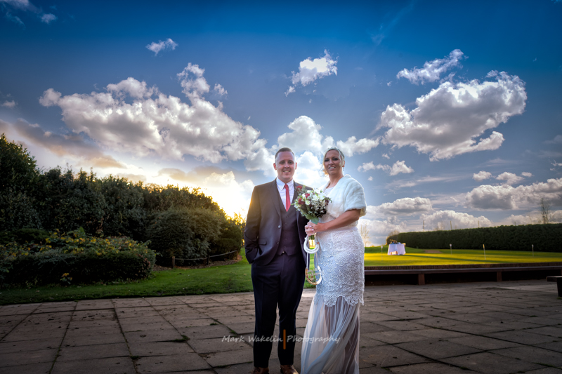 Bride and groom standing outdoors on a paved patio with a blue sky and scattered clouds at sunset.