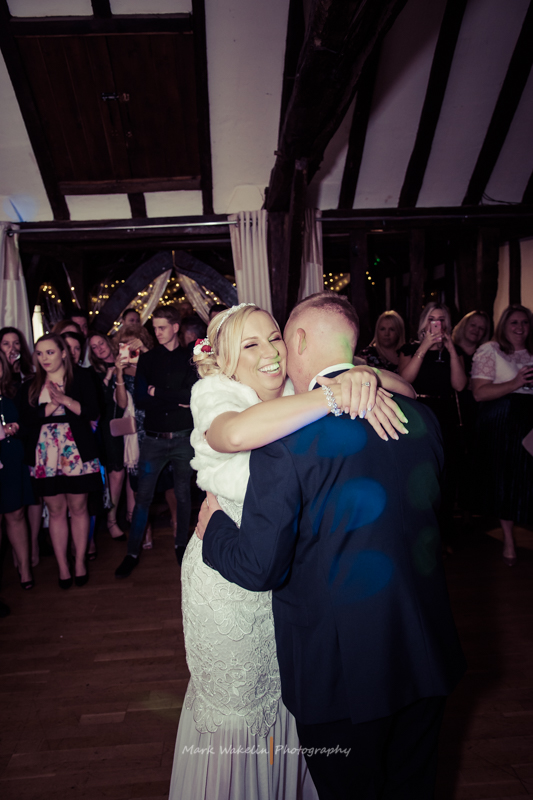 Bride and groom dancing closely and smiling in a warmly lit venue with guests watching.