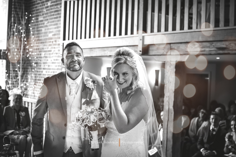 Bride and groom standing together indoors, the bride smiling and holding a bouquet while wiping a tear.