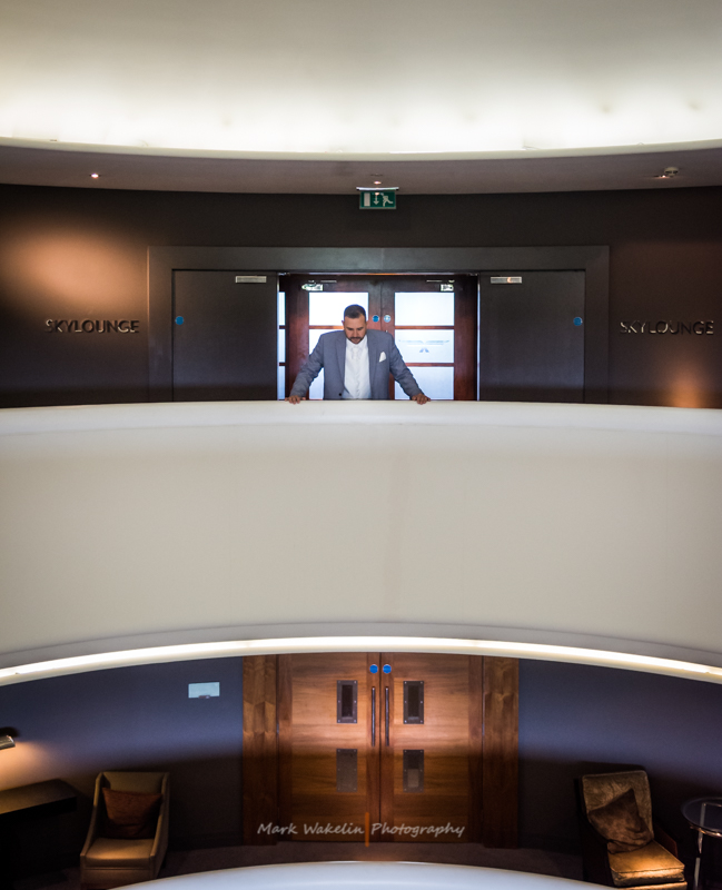 Man in a suit leaning on a white curved balcony railing inside a stylish lounge with wooden double doors and dim lighting.
