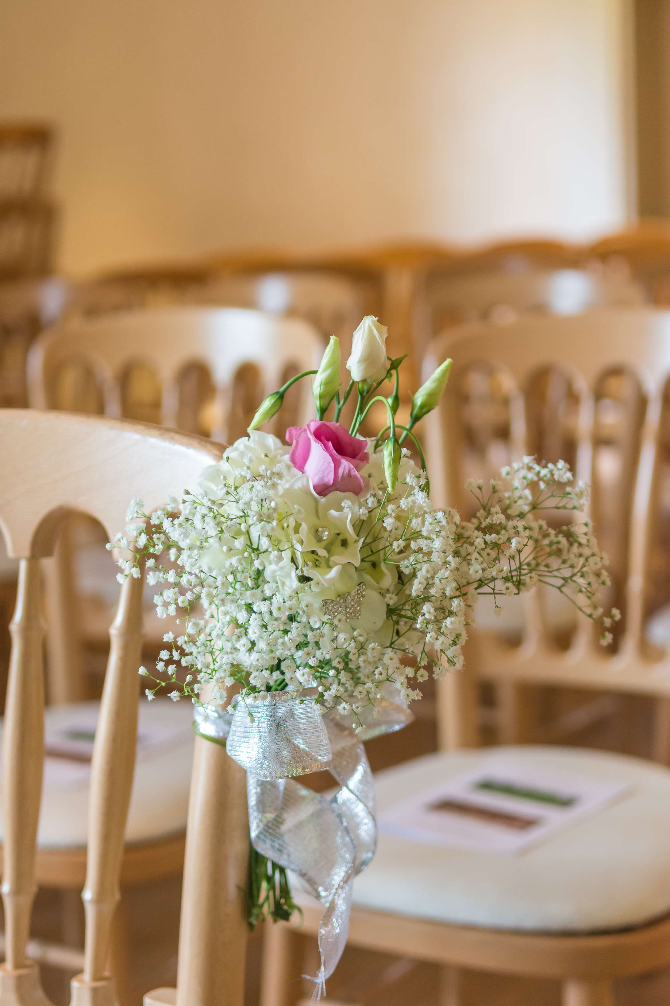 Small bouquet of white and pink flowers tied with silver ribbon attached to a wooden chair at a wedding venue.