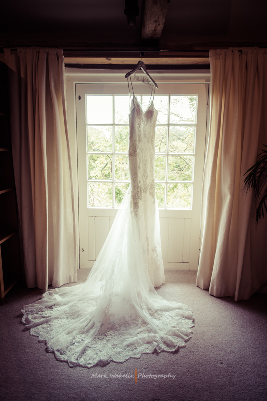 White lace wedding dress hanging in front of a window with light curtains.