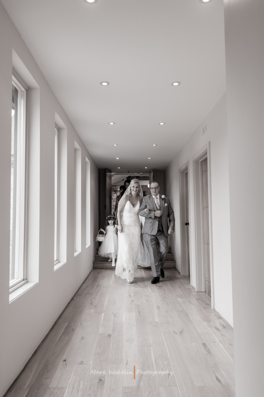 Bride in a lace wedding dress walking arm-in-arm down a hallway with a man in a suit, followed by a flower girl holding a basket.