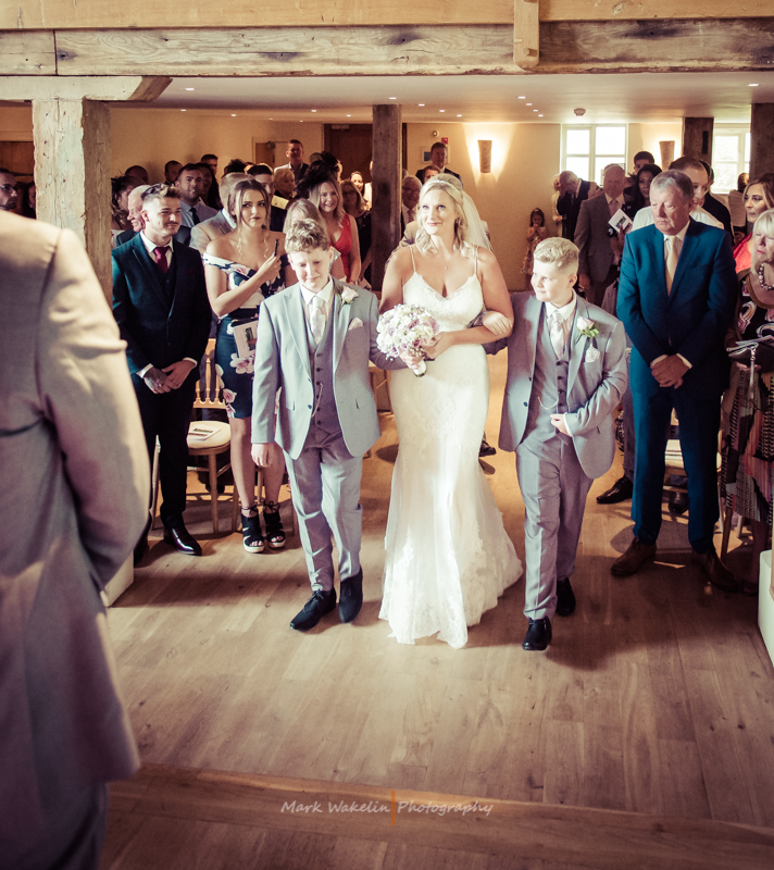Bride in white wedding dress holding a bouquet walking down the aisle indoors, flanked by two boys in light grey suits, with guests watching in the background.