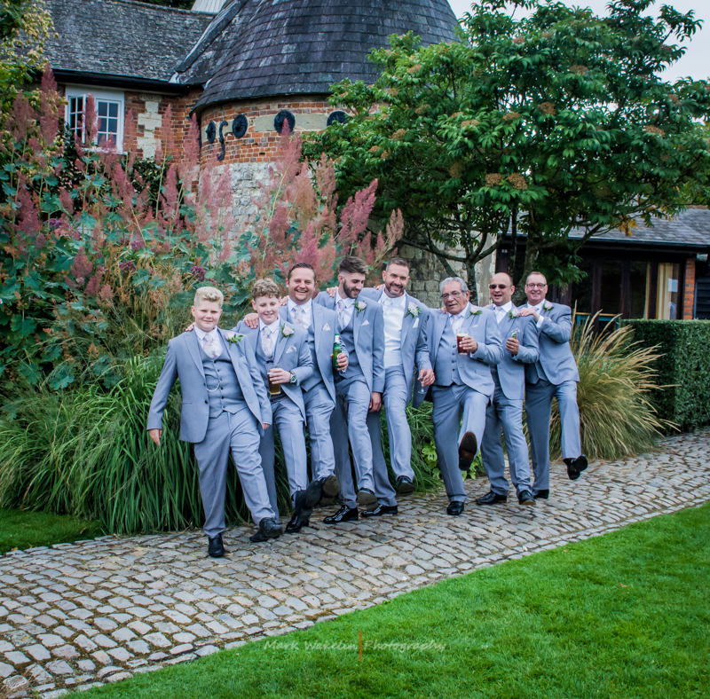 Seven men dressed in light grey suits with boutonnieres stand on a cobblestone path, smiling and kicking their legs in front of a garden with tall plants and a stone building.
