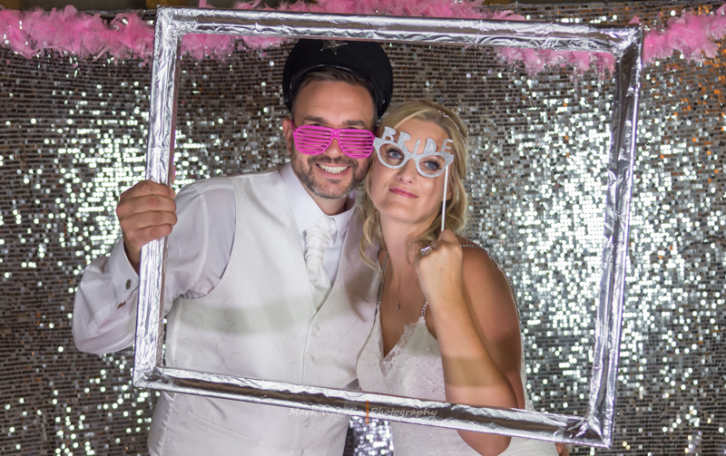 Smiling bride and groom posing with fun props inside a silver picture frame at a wedding photo booth.