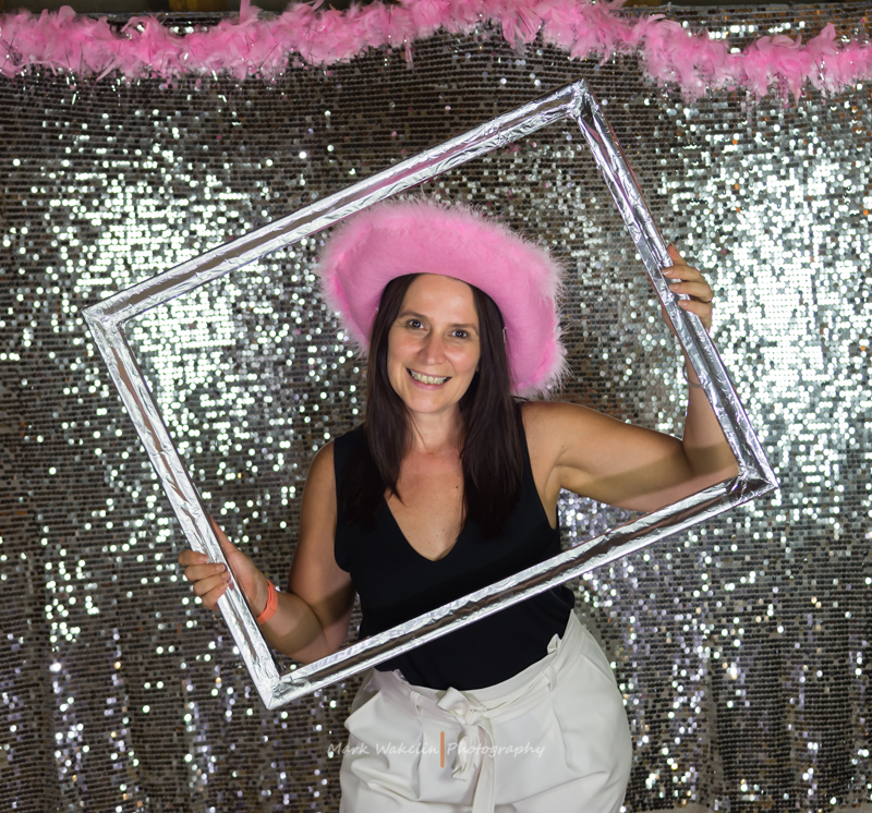 Smiling woman in a pink fuzzy hat holding a silver picture frame against a sparkly sequin backdrop with a pink feather decoration above.
