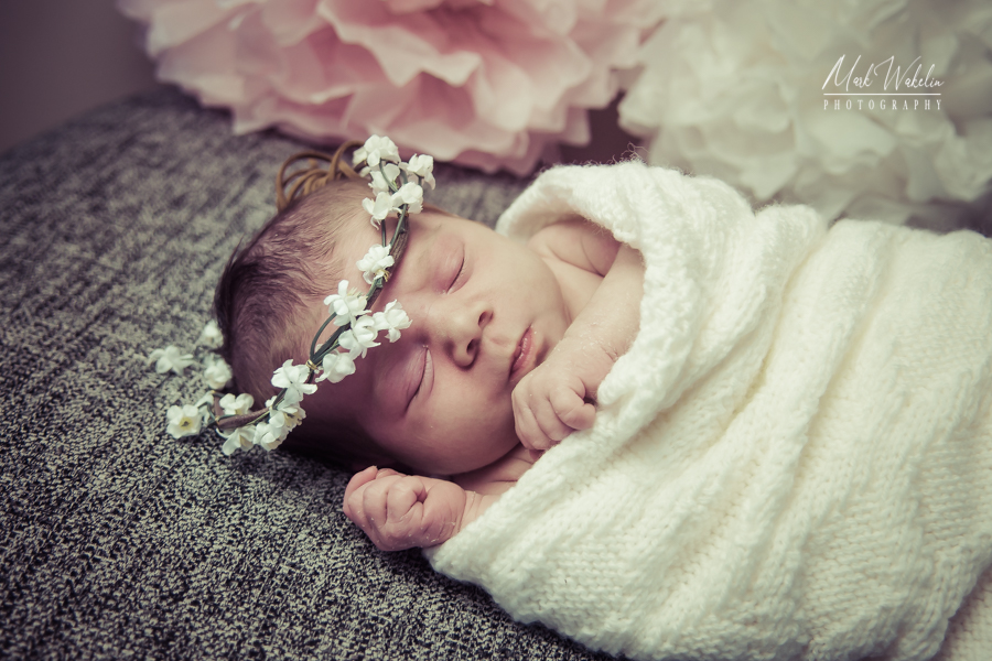 Newborn baby sleeping peacefully wrapped in a white knitted blanket with a white flower crown on their head.
