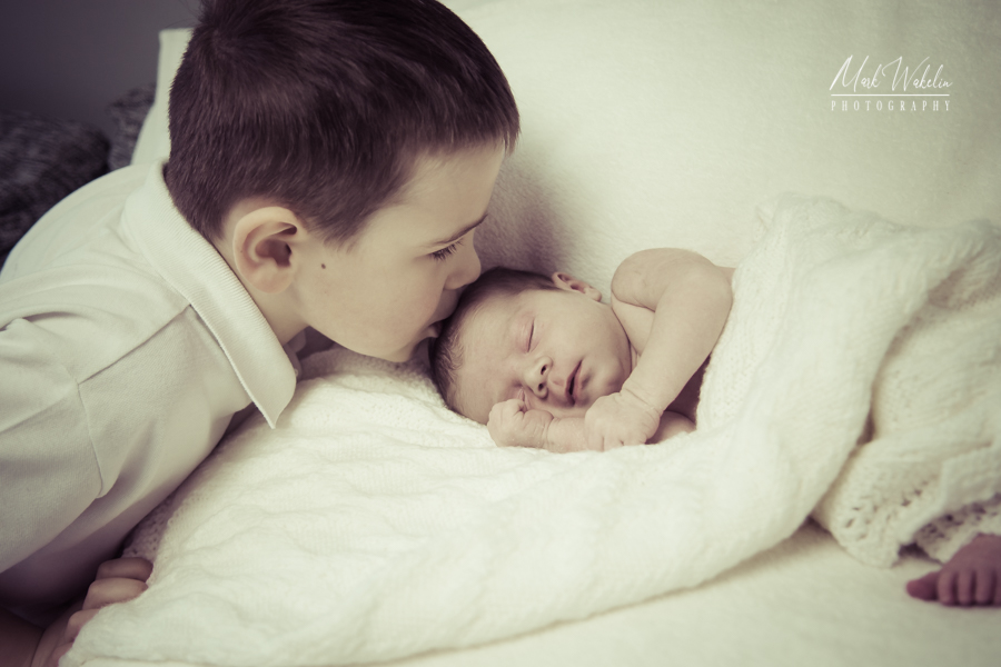Young boy gently kissing a sleeping newborn baby wrapped in a white knitted blanket.