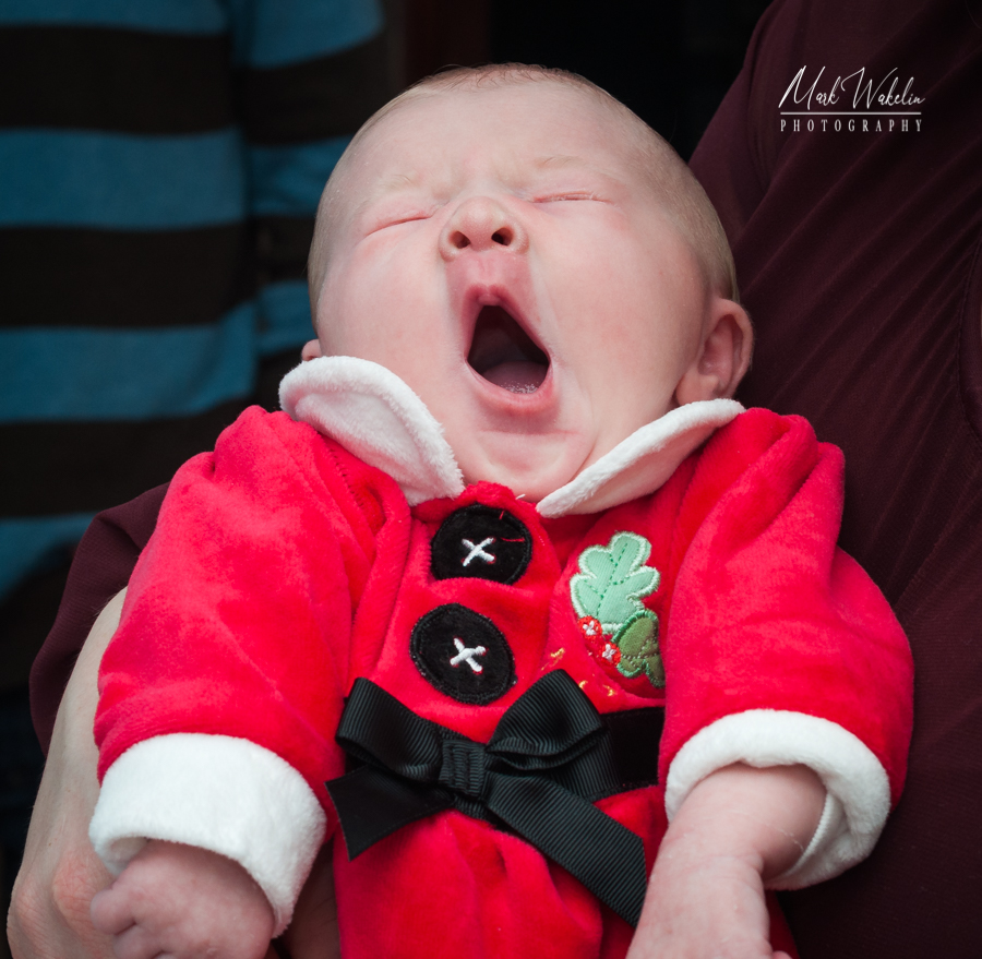 Yawning baby wearing a festive red outfit with white trim and black bow being held.