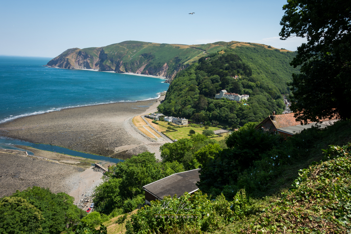 Coastal view of a rocky beach with green hills and scattered houses under a clear blue sky.