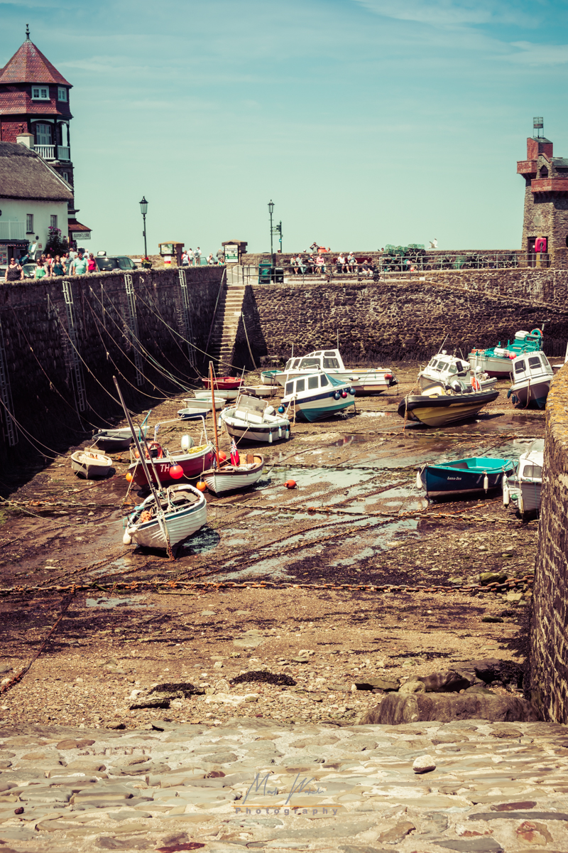 Boats resting on a dry dock at low tide within a stone harbour surrounded by old buildings and people walking along the pier under a clear sky.