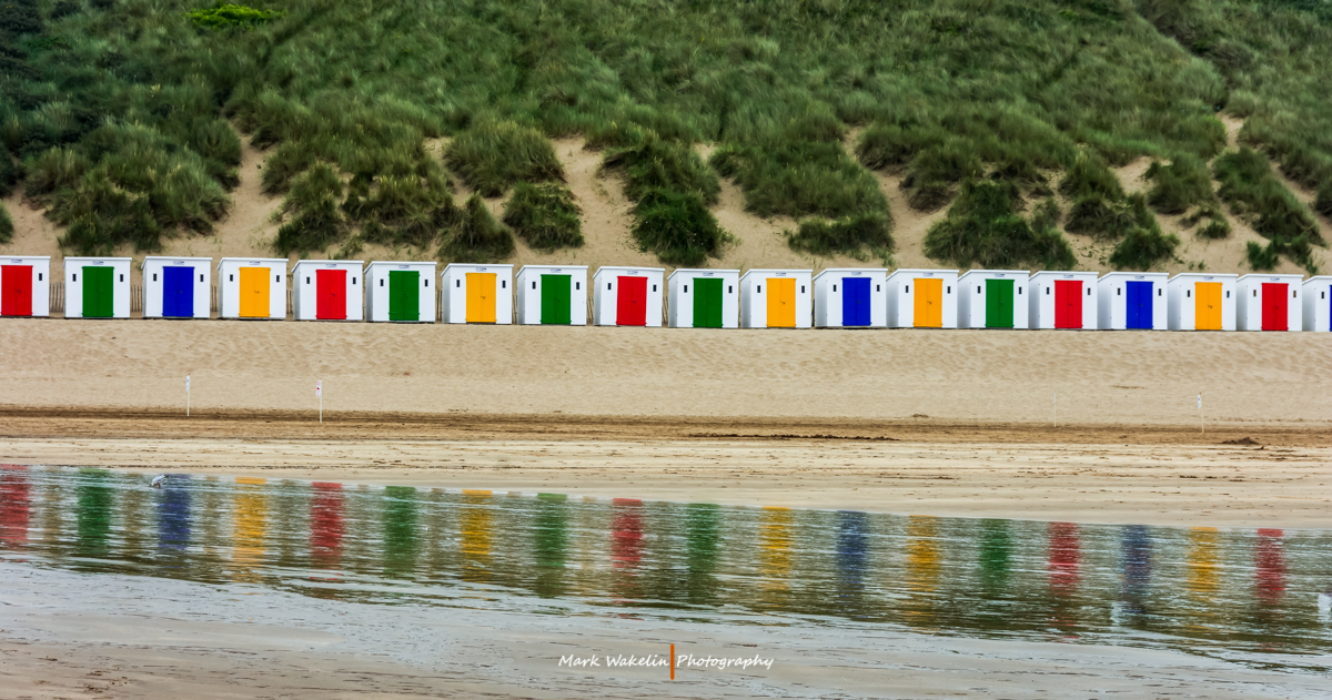 Row of white beach huts with red, green, blue and yellow doors reflected in wet sand, backed by grassy dunes.
