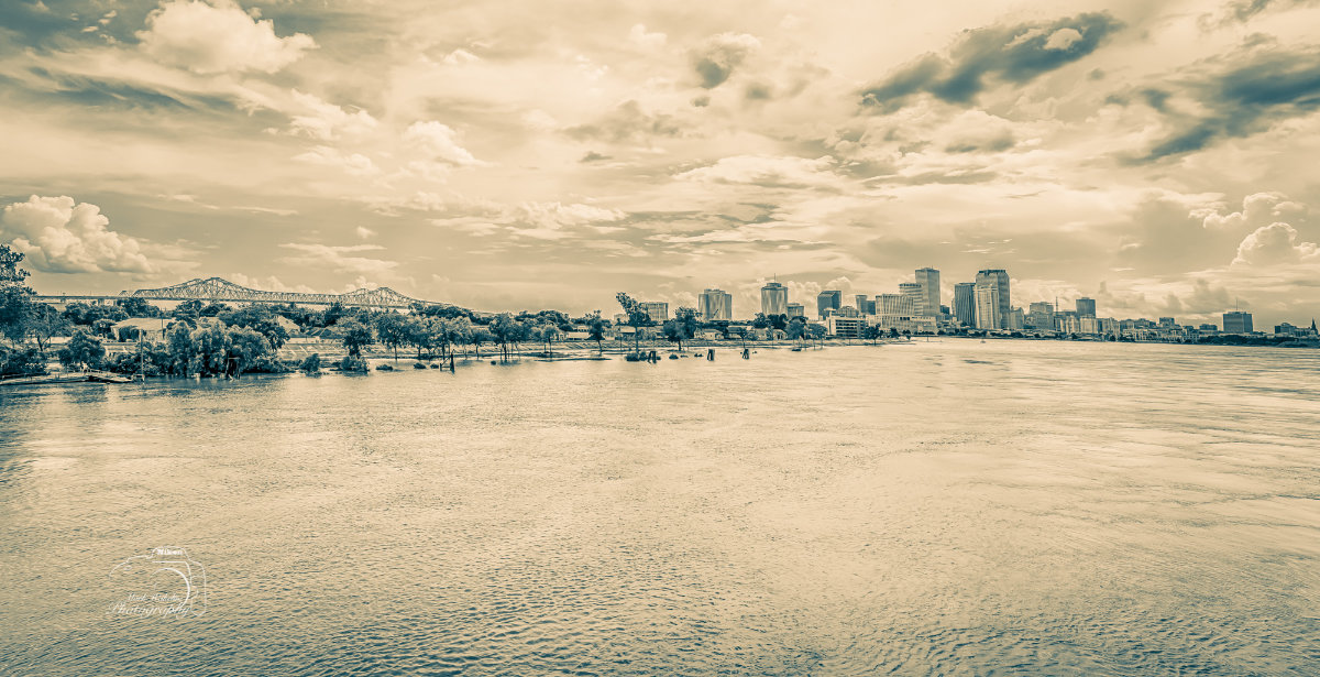 Panoramic view of a city skyline behind a river with a bridge and trees under a cloudy sky.