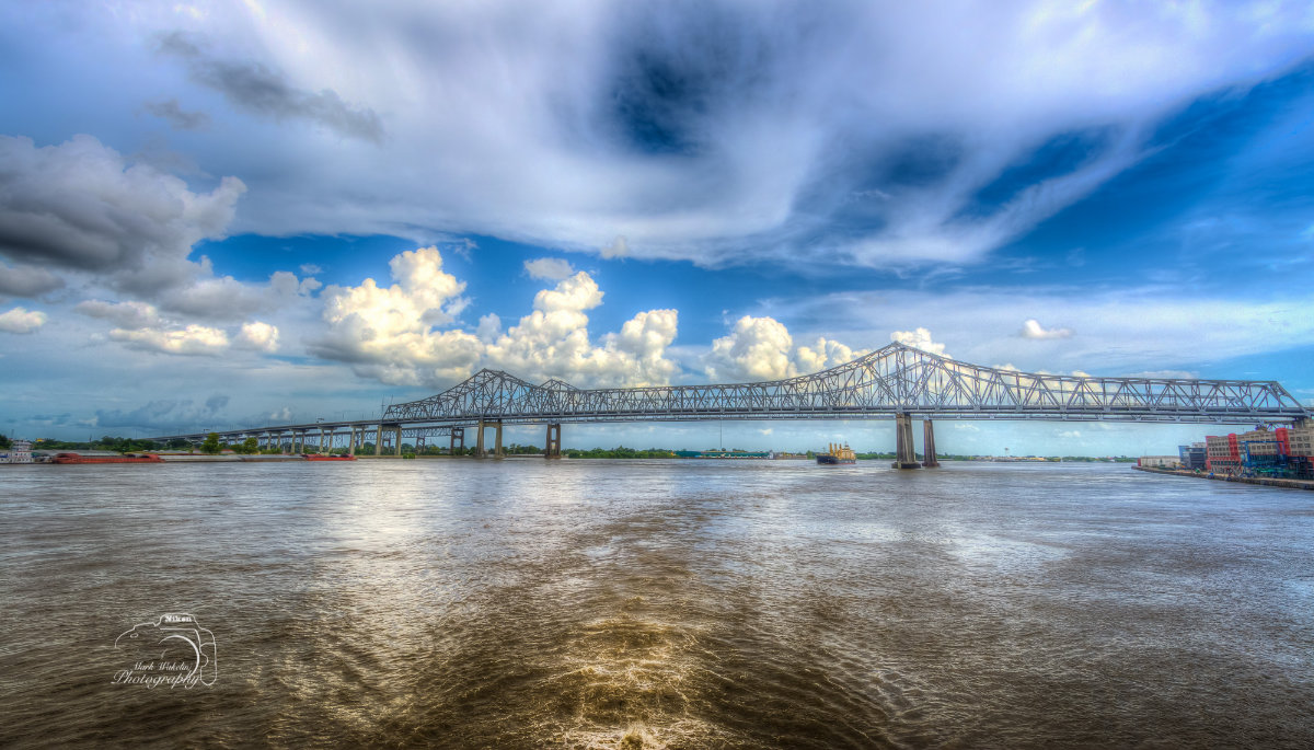 Long steel truss bridge spanning a wide river under a dramatic cloudy blue sky.