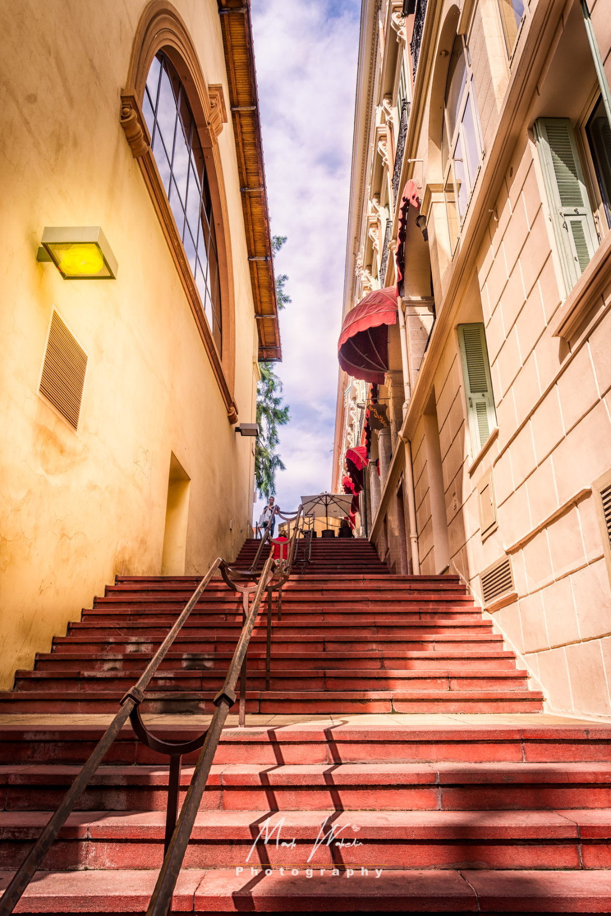 Wide red staircase flanked by beige buildings with awnings under a partly cloudy sky.