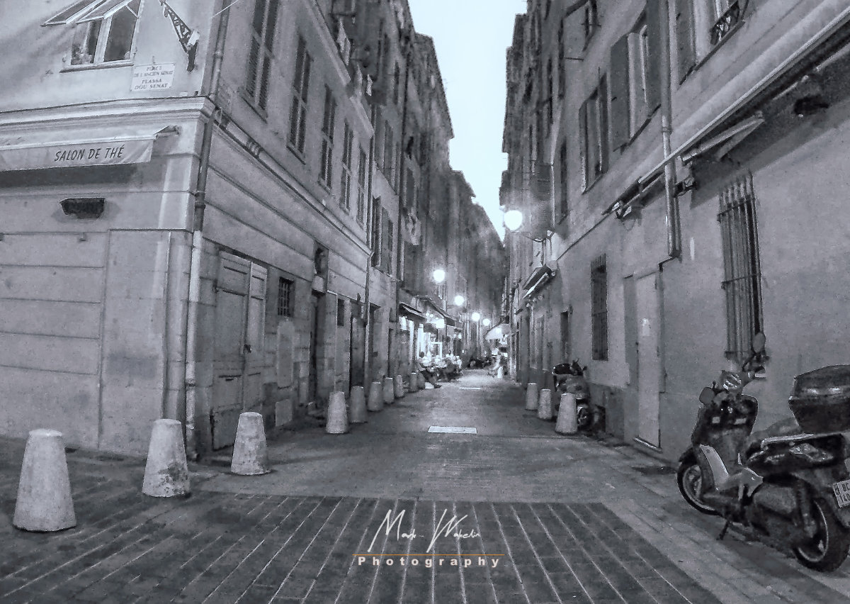 Narrow cobblestone street in a European city at dusk with scooters parked on the right and streetlights illuminating the path.