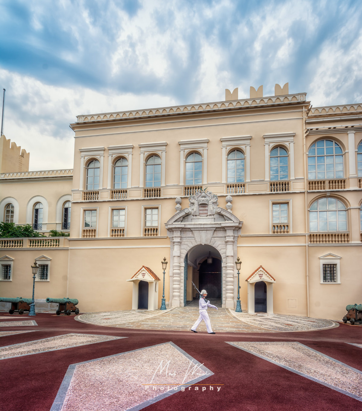 Guard in white uniform marching in front of a beige historic palace entrance under a cloudy sky.