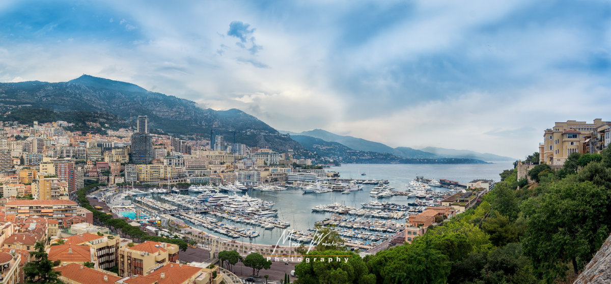 Panoramic view of a coastal city with a marina filled with yachts, surrounded by buildings and green hills under a partly cloudy sky.