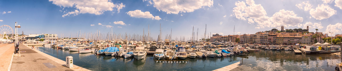 Panoramic view of a marina filled with boats under a partly cloudy sky next to a coastal town with colourful buildings.