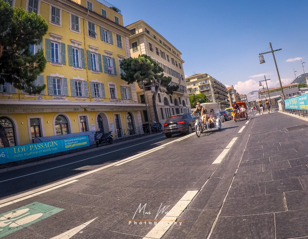 Street scene with people riding rickshaws and a black car parked by yellow and beige buildings under a clear blue sky.