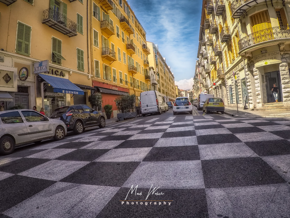 Urban street with a distinctive black and white checkerboard pattern painted on the road, lined with parked cars and yellow buildings under a partly cloudy sky.