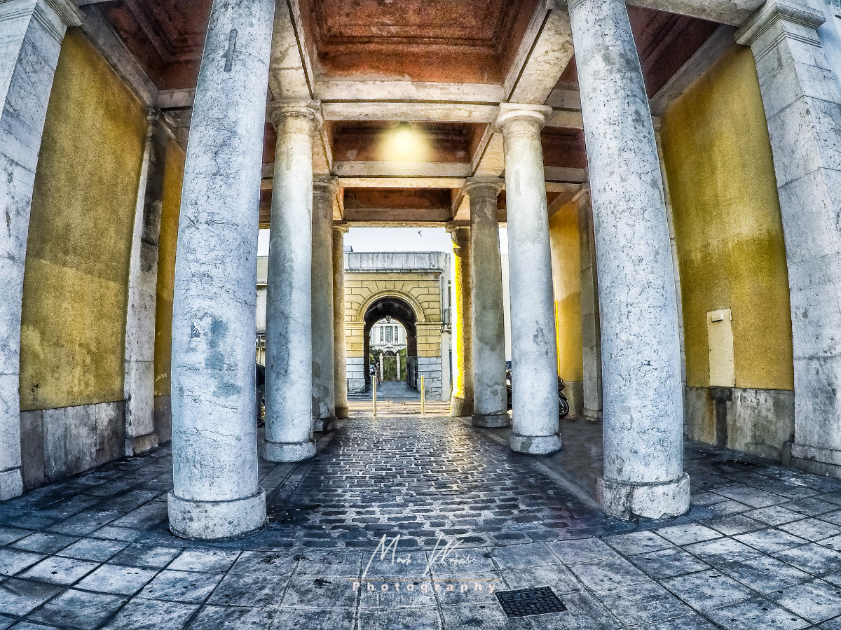 Stone archway with tall marble columns and yellow walls leading to a cobblestone path and another arch in the background.