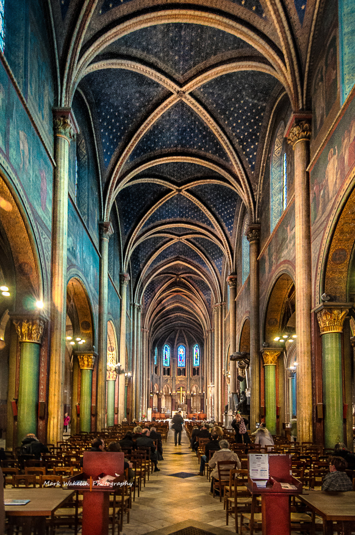 Interior of a large cathedral with vaulted ceilings, stained glass windows, and rows of wooden chairs filled with people.