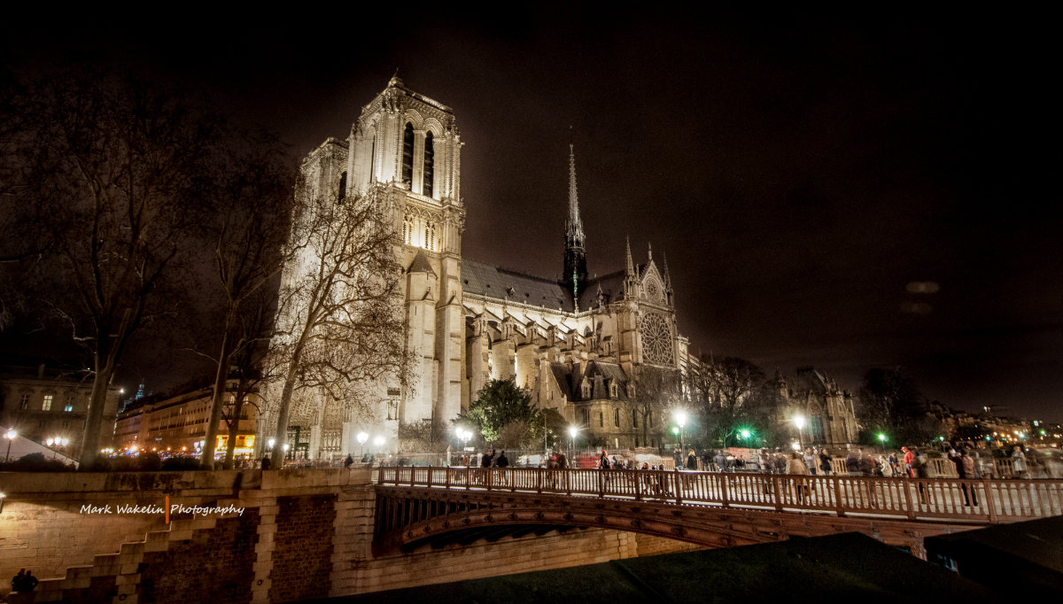 Notre-Dame Cathedral in Paris illuminated at night with people walking on the nearby bridge.