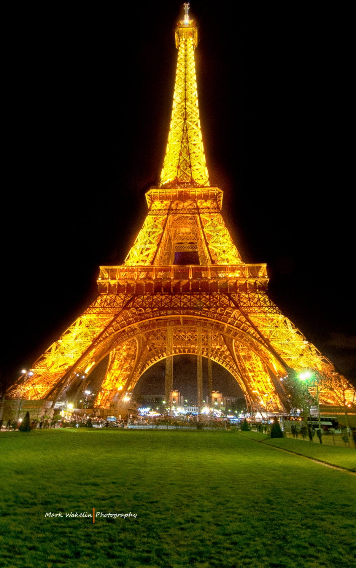 Eiffel Tower illuminated with golden lights at night over green lawn with people around.