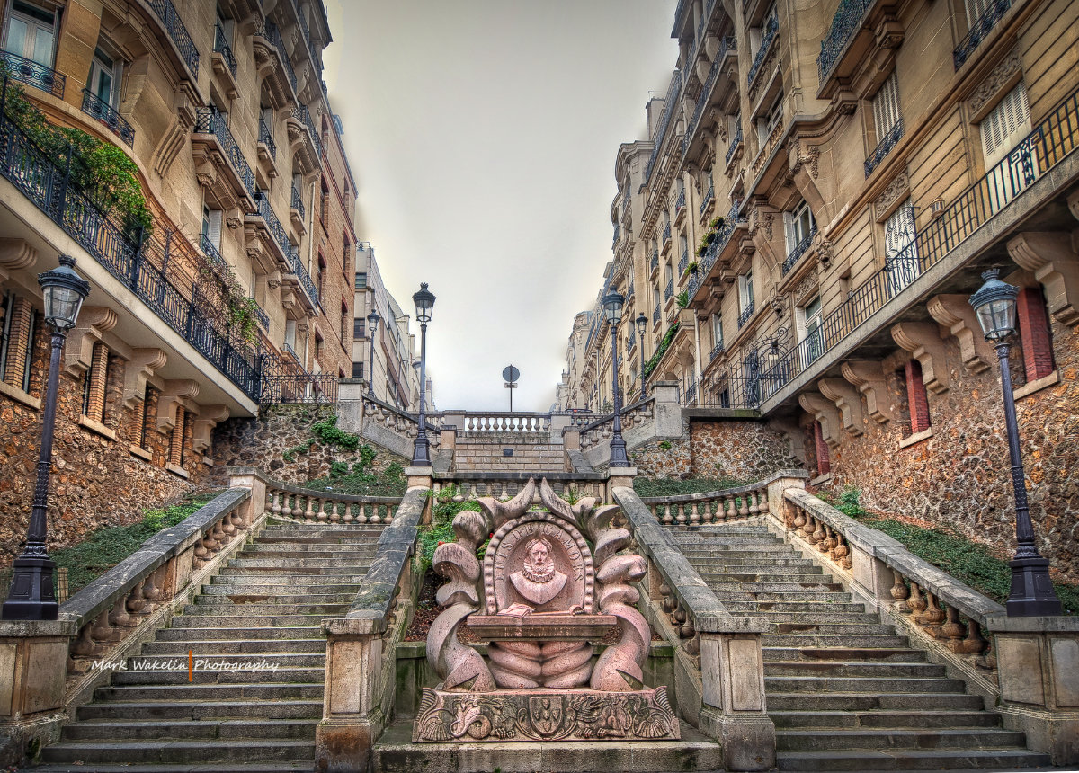 Symmetrical stone staircase with ornate balustrades flanked by historic Parisian buildings and a central sculpted fountain featuring a bust.