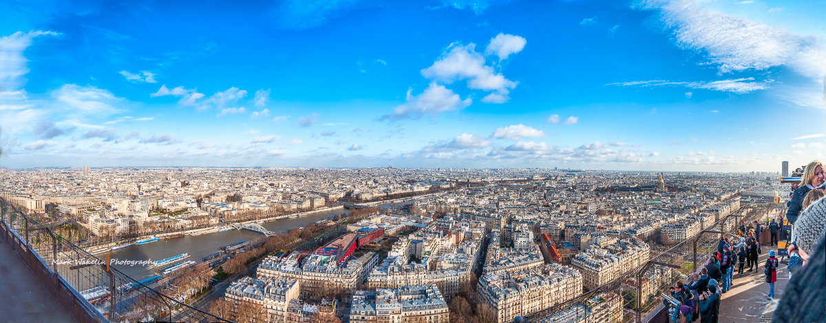 Panoramic view of Paris with the River Seine, historic buildings, and blue sky with scattered clouds, seen from a high vantage point crowded with tourists.
