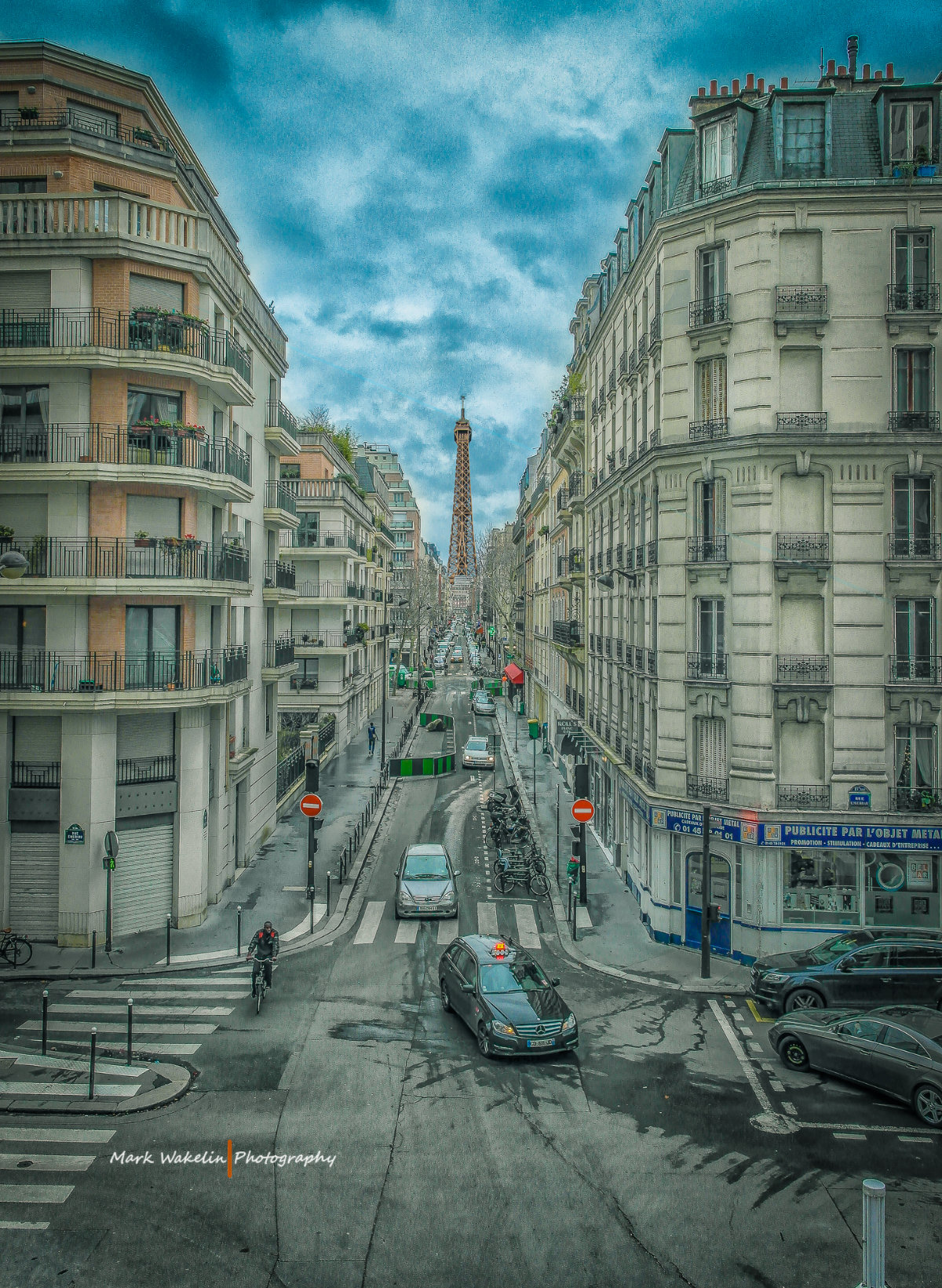 View down a Paris street with apartment buildings on both sides and the Eiffel Tower in the distance under a cloudy sky.