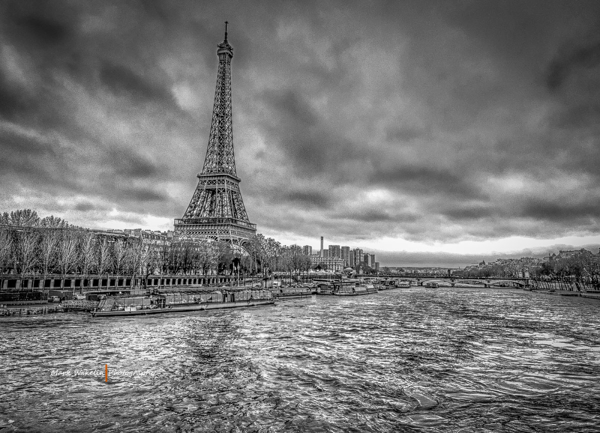 Black and white image of the Eiffel Tower beside the River Seine with boats and cloudy sky in Paris.