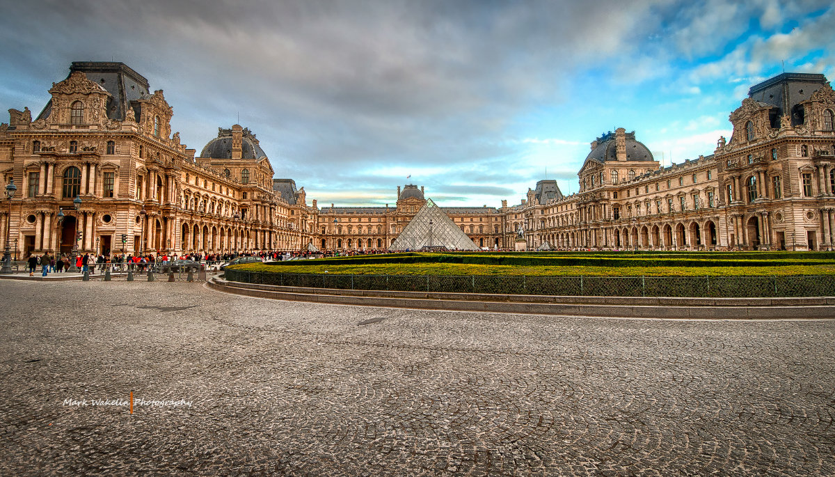 Wide view of the Louvre Museum with its glass pyramid entrance under a partly cloudy sky.