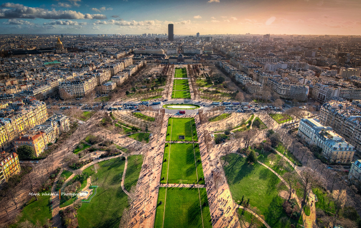 Aerial view of a large park with green lawns, tree-lined paths, and busy city streets under a partly cloudy sky at sunset.