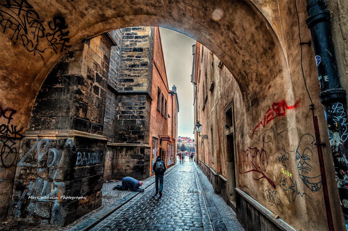 Narrow cobblestone street framed by an arch with graffiti, featuring a person walking and another kneeling on the ground.