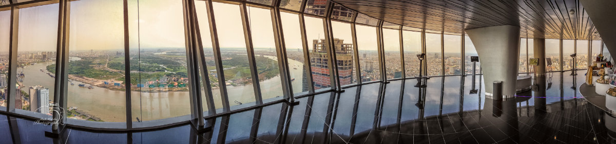 Panoramic indoor view from a high-rise building overlooking a river with boats and a sprawling cityscape under warm sunlight.