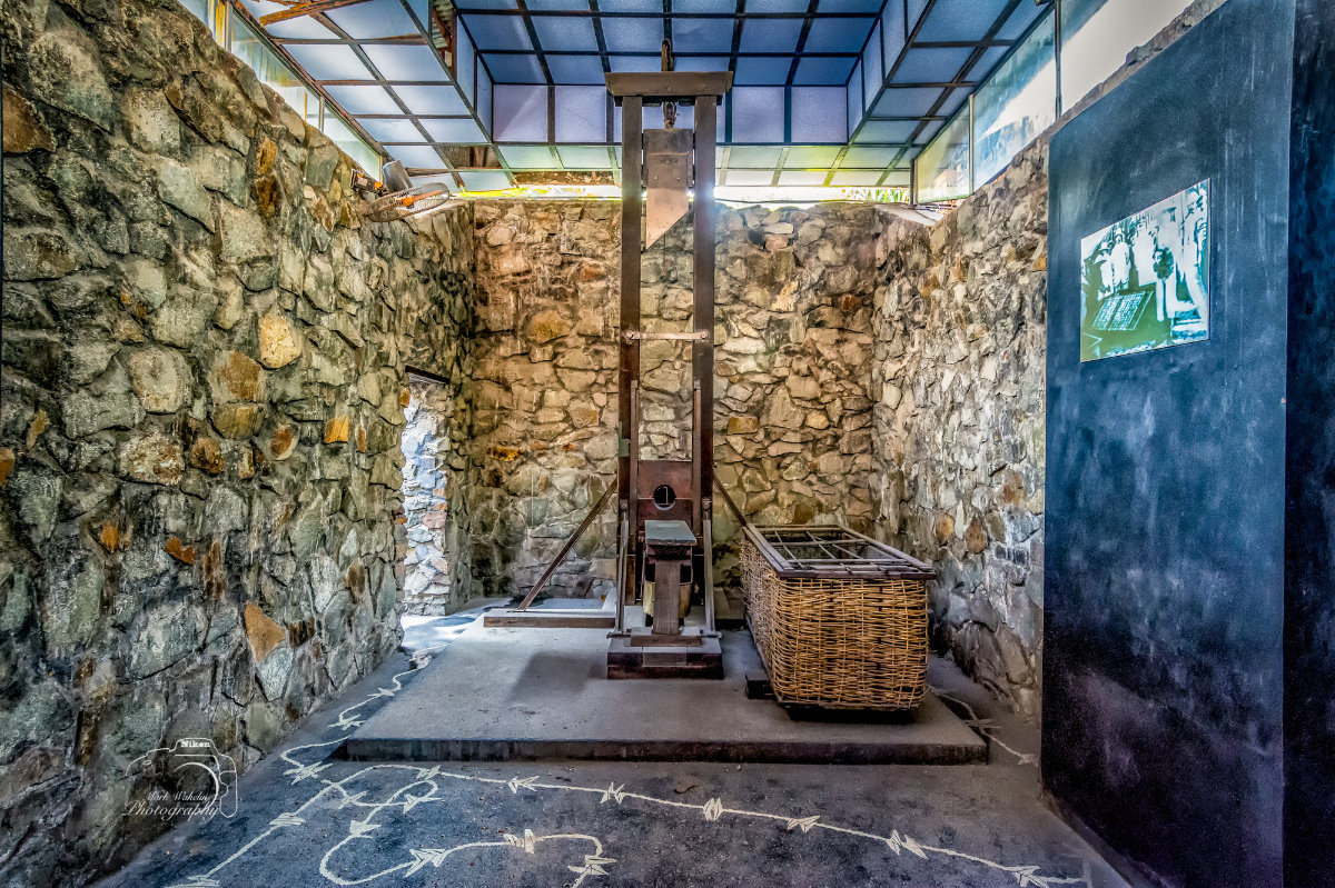 Stone-walled room with a guillotine and a wicker basket, featuring white chalk arrows on the floor showing a path.