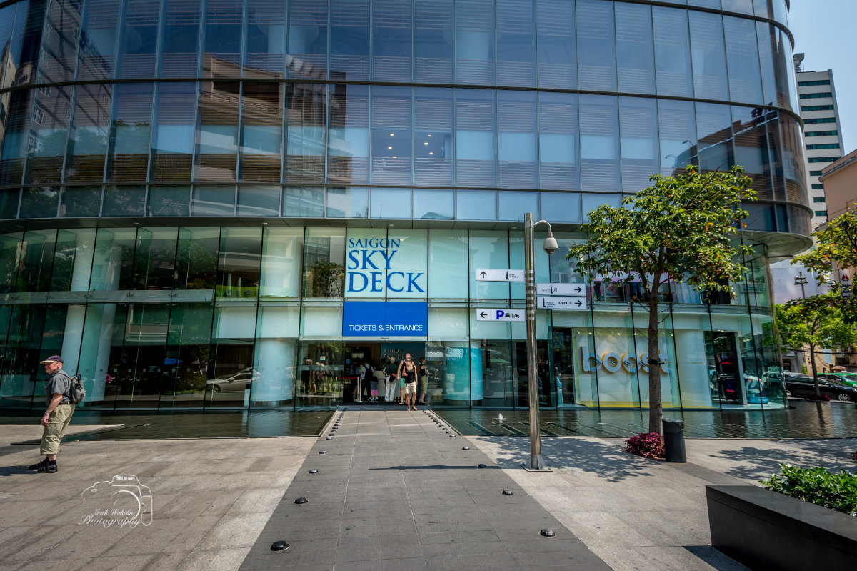 Entrance to the Saigon Sky Deck with people walking on a path leading to the ticket and entrance area beneath a modern glass building.
