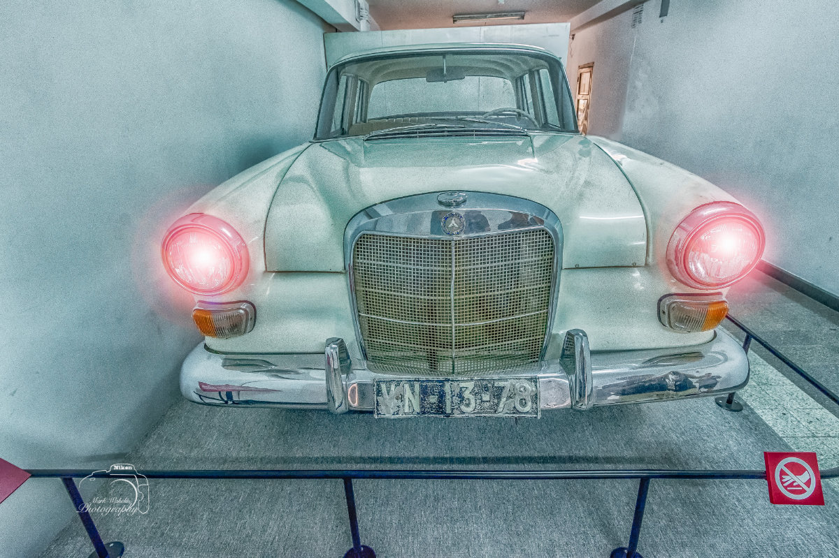 Front view of a vintage Mercedes-Benz car with headlights on, displayed indoors behind a barrier.
