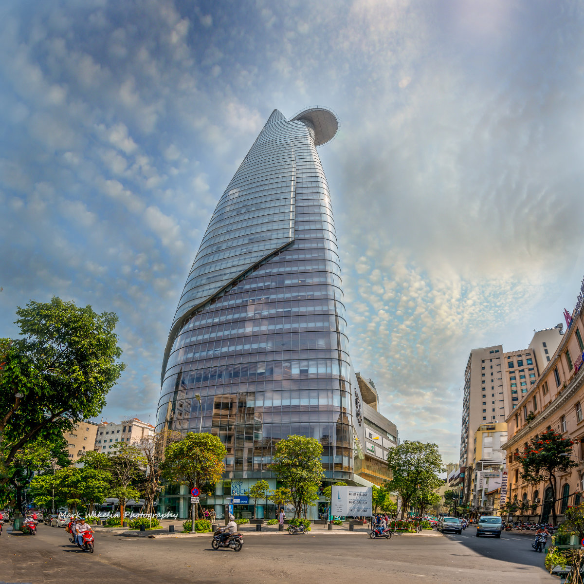 Modern curved glass skyscraper with observation deck towering over a busy street with motorbikes and trees under a partly cloudy sky.